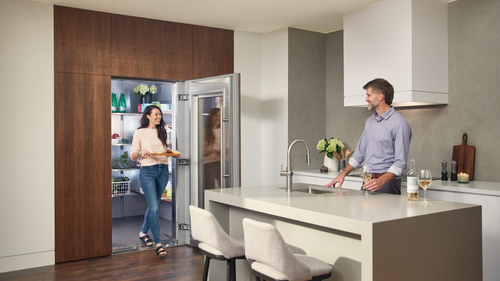 A woman carrying a tray of food walks out of a Qoldfusion walk-in refrigerator while a man stands at a kitchen island smiling, with wine glasses on the counter. The kitchen features a minimalist design with wooden cabinetry and a light-colored countertop.