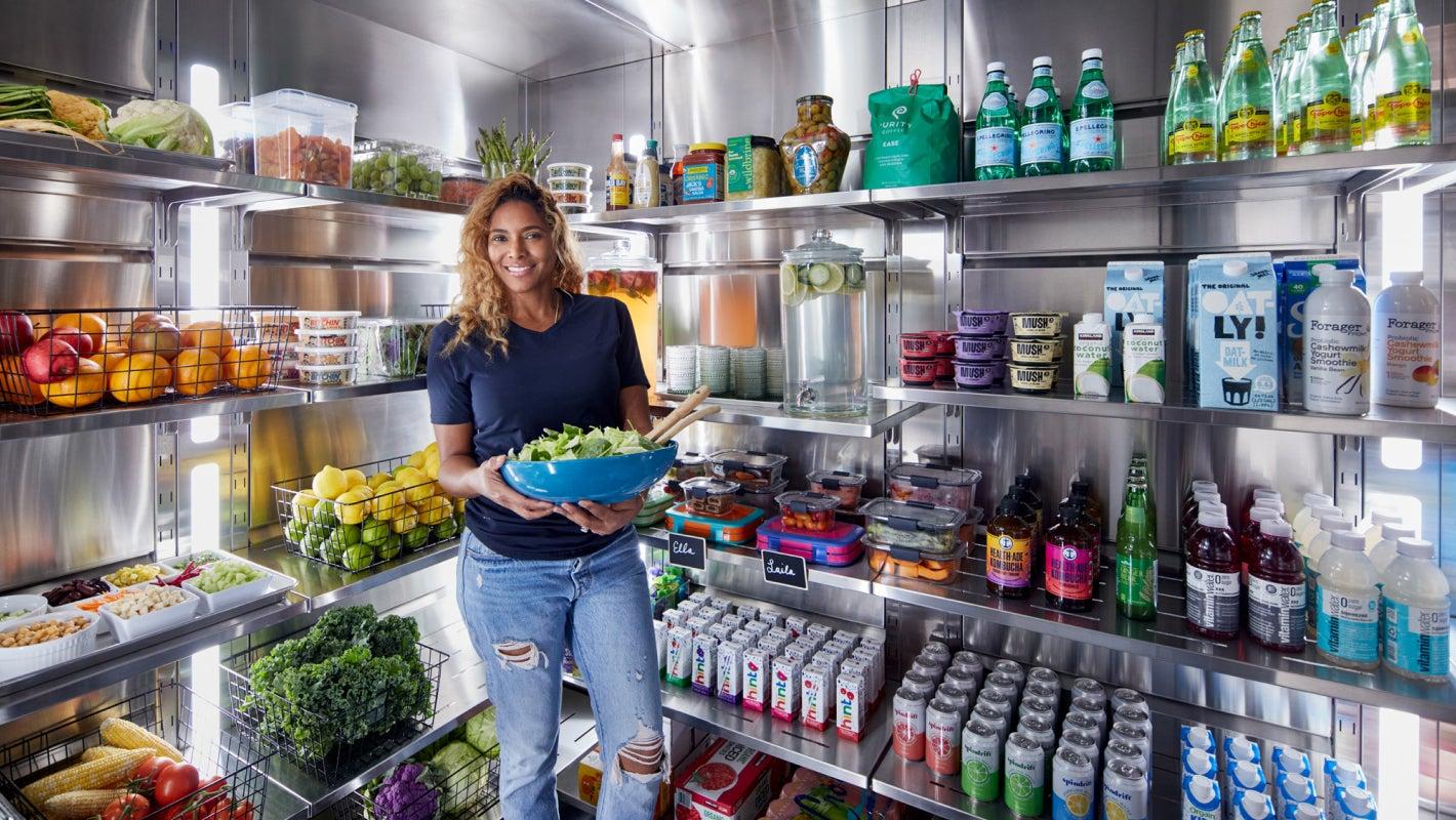 A woman holding a bowl of salad stands inside a well-organized luxury Qoldfusion walk-in cold pantry filled with various fresh produce, beverages and more.