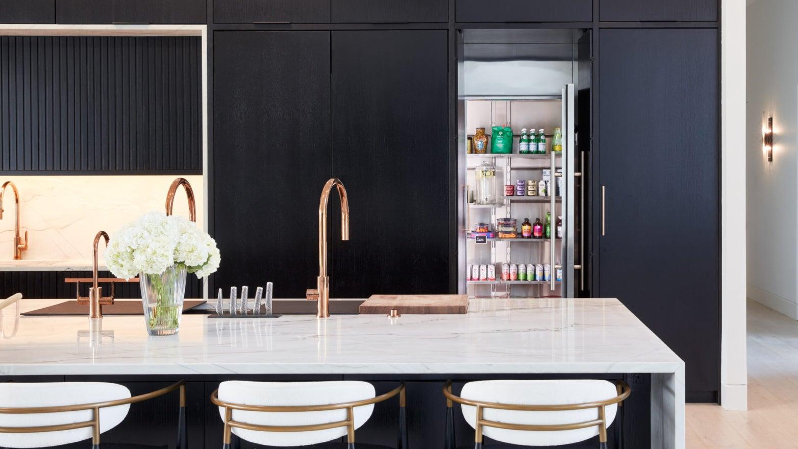 Modern kitchen with black cabinetry, marble countertop, and a vase of white flowers. A Qoldfusion walk-in refrigerator is visible with organized food items.